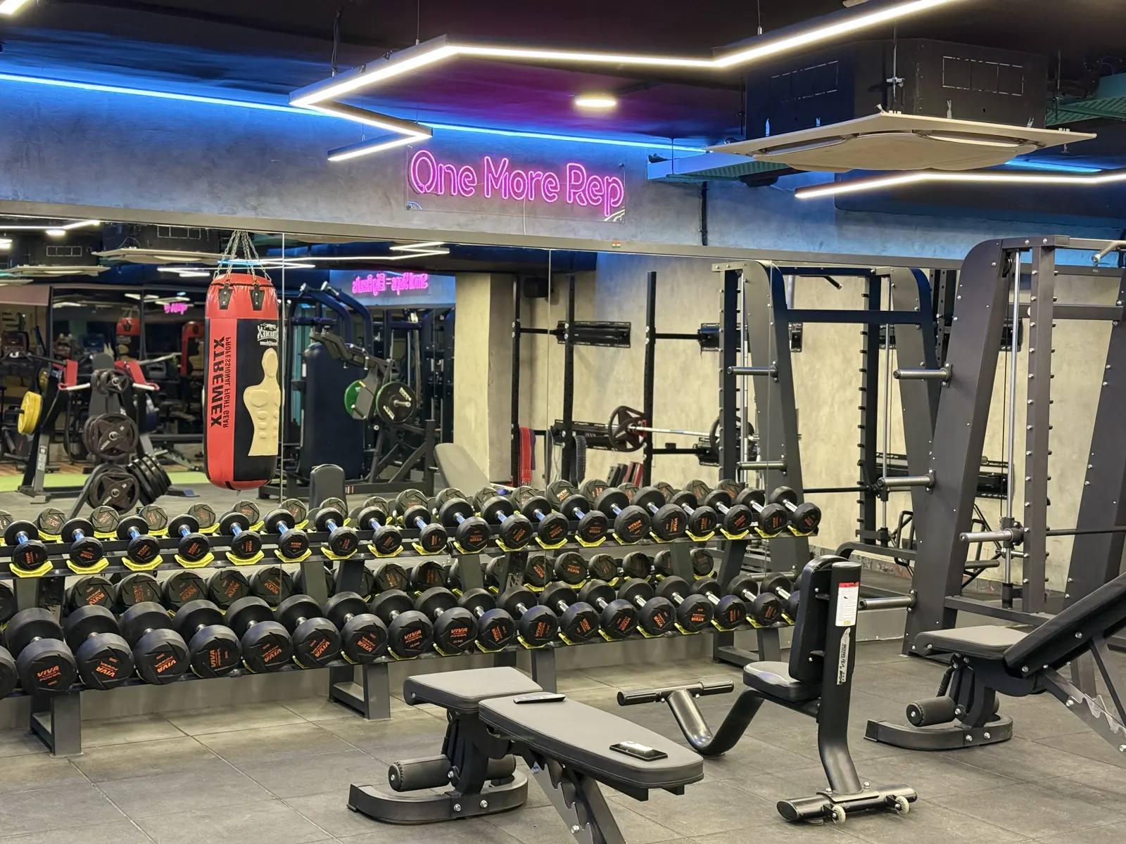 The Fisique floor: a full dumbbell rack and benches under the pink neon One More Rep sign